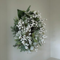 large artificial Christmas wreath made up with a full arrangement of mixed foliages in soft, muted green tones, dressed with delicate snowberries and lightly dusted with frosted snow and iridescent glitter for a subtle sparkle